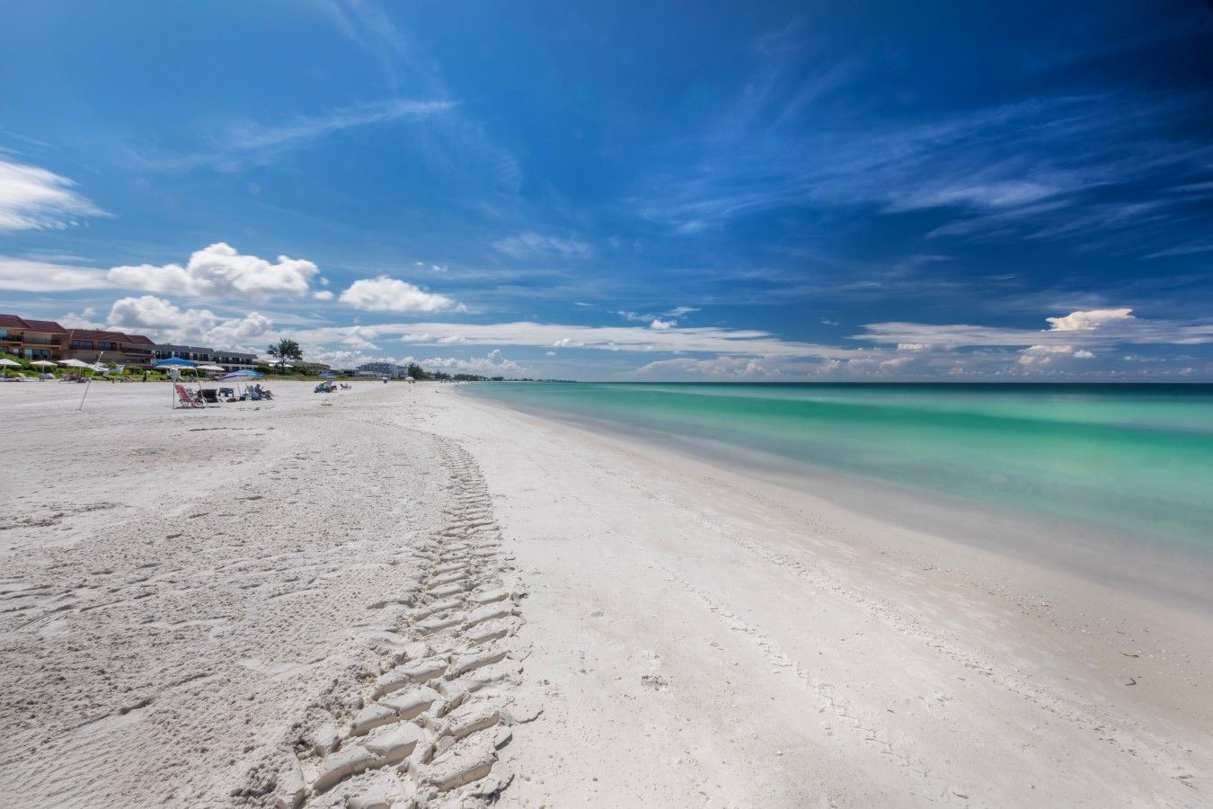 A beach with a clear sky, a sandy shore, and a few buildings in the background.