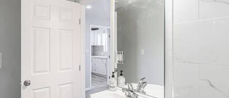 A bathroom with a white sink, a mirror, and a door leading to a kitchen area.
