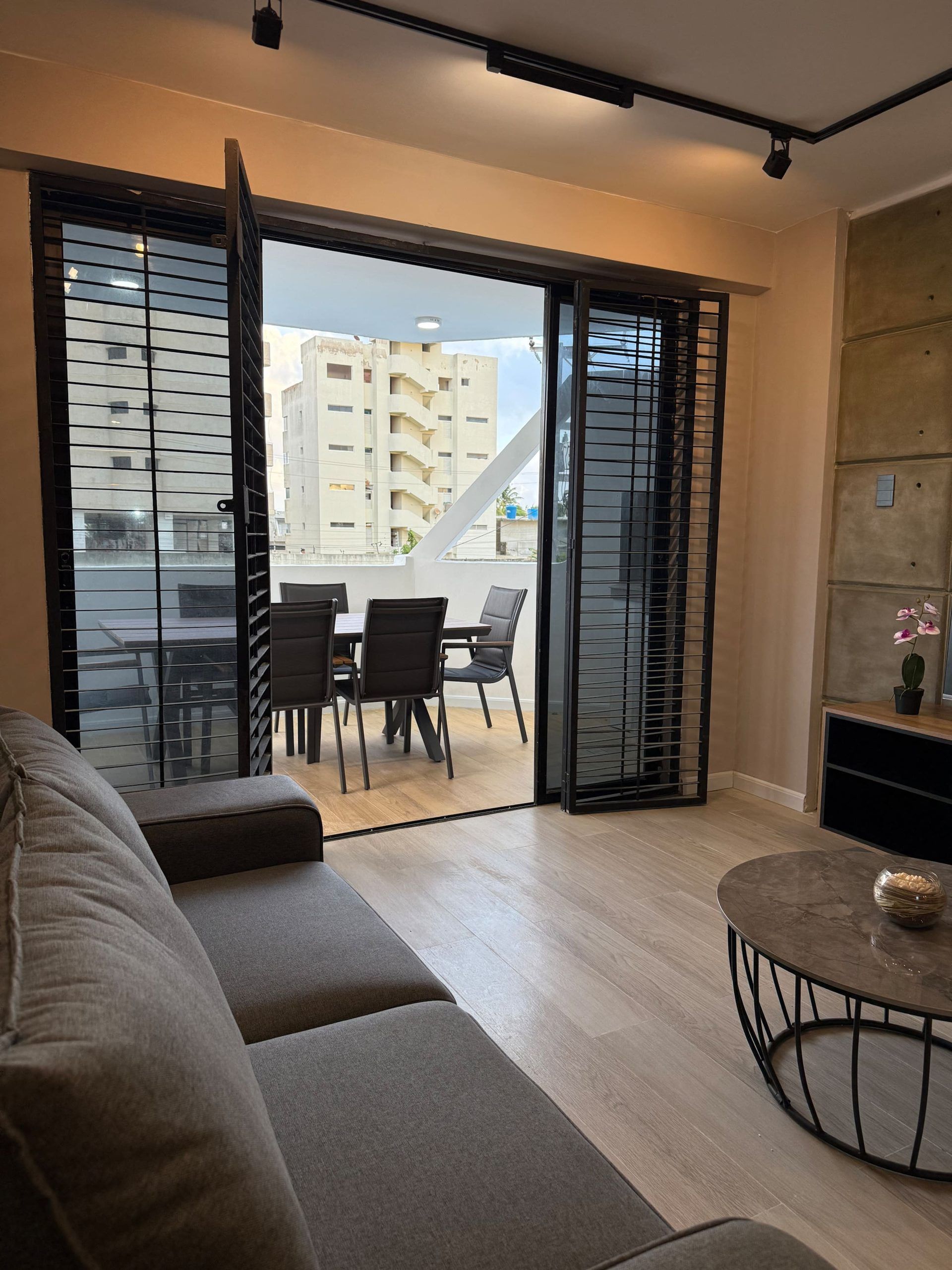 A modern living room with a sofa, a dining table with chairs, and a balcony with a view of buildings.