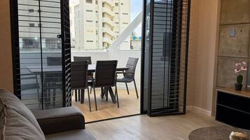 A modern living room with a sofa, a dining table with chairs, and a balcony with a view of buildings.