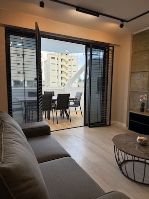 A modern living room with a sofa, a dining table with chairs, and a balcony with a view of buildings.