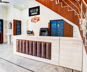 A hotel reception desk with a sign above it, a staircase to the right, and a wall-mounted printer. - Saver Double Hotel White Retreat Inn (Vikasnagar)
