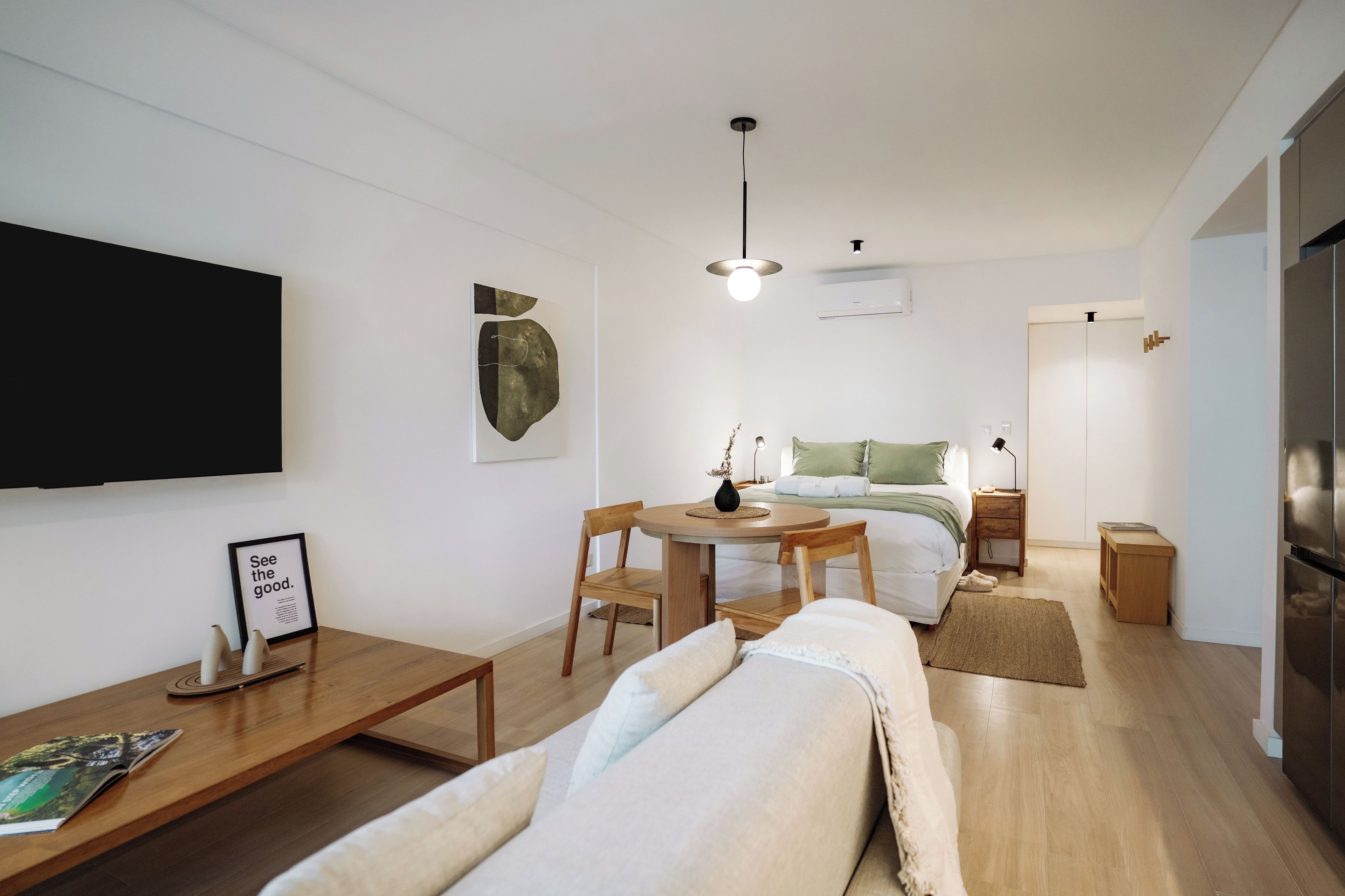 A modern, well-lit living room with a flat-screen TV, a dining area with a table and chairs, a bed with a green headboard, and a kitchen area with a refrigerator.