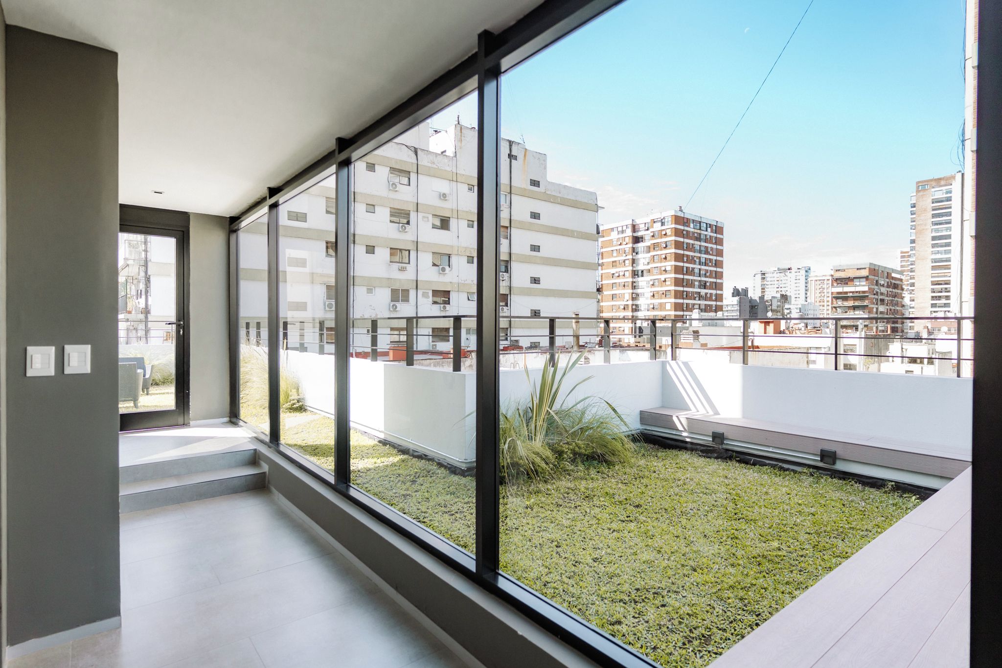 Un apartamento moderno con ventanas amplias, un balcón con césped y vistas a edificios circundantes.