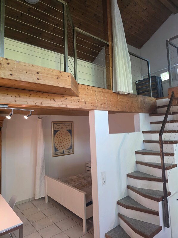 A bedroom with a wooden ceiling, a bed, a staircase, and a window.