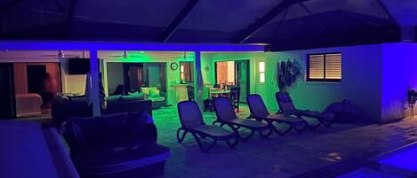 A room with a pool, lounge chairs, and a dining area.