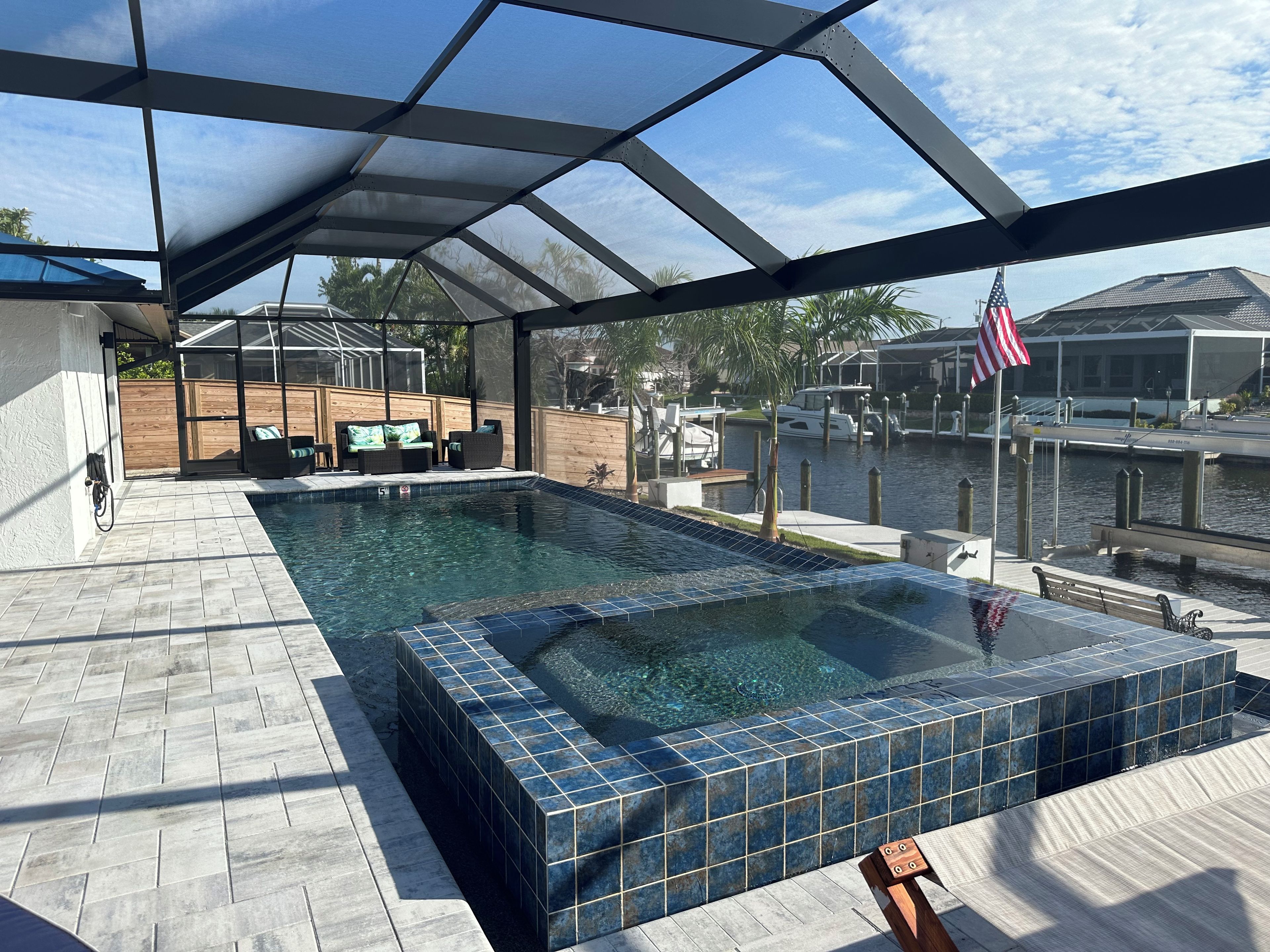 Ein überdachter Außenpool mit Whirlpool, einer Holzterrasse und Blick auf einen Yachthafen mit Booten und einer amerikanischen Flagge.