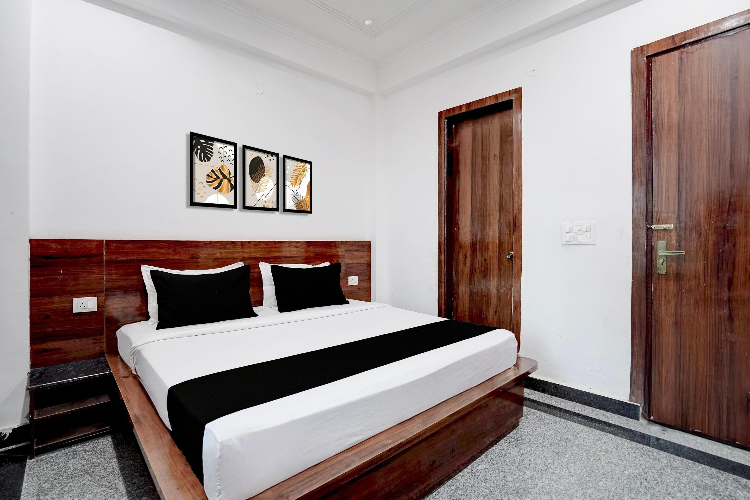 Quarto com cabeceira de madeira, cama com roupa de cama branca, almofadas pretas e faixa decorativa preta. Três obras de arte emolduradas estão penduradas na parede acima da cama.
