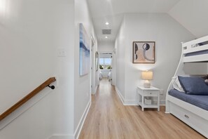 A narrow hallway with a wooden handrail, a white bedside table with a lamp, and a bunk bed with blue bedding.