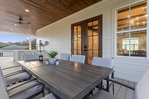 Un espacio exterior techado para comer con una mesa y sillas de madera, plantas en macetas y vista de la casa.