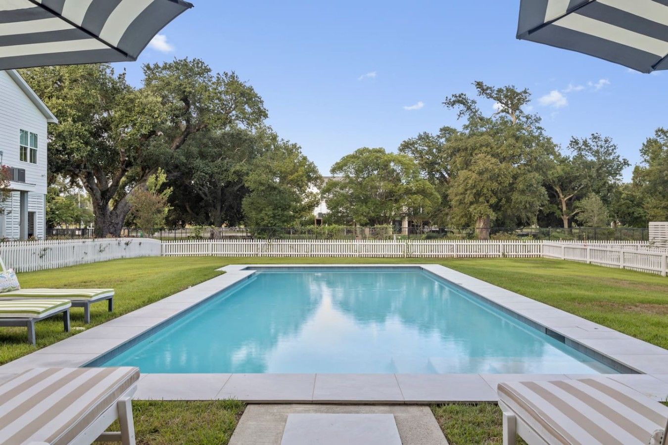Et rektangulært swimmingpool omgivet af et hvidt hegn, to liggestole og et hvidt stakhegn i baggrunden.