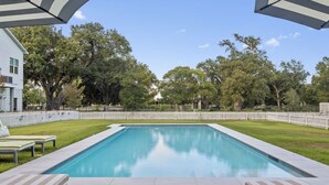Una piscina rectangular con una cerca blanca, dos sillas de descanso y una valla de piquetes blancos al fondo.