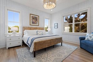 A bedroom with a large bed, two bedside tables, a dresser, and a blue sofa. There are two windows with views of trees and a ceiling light fixture.