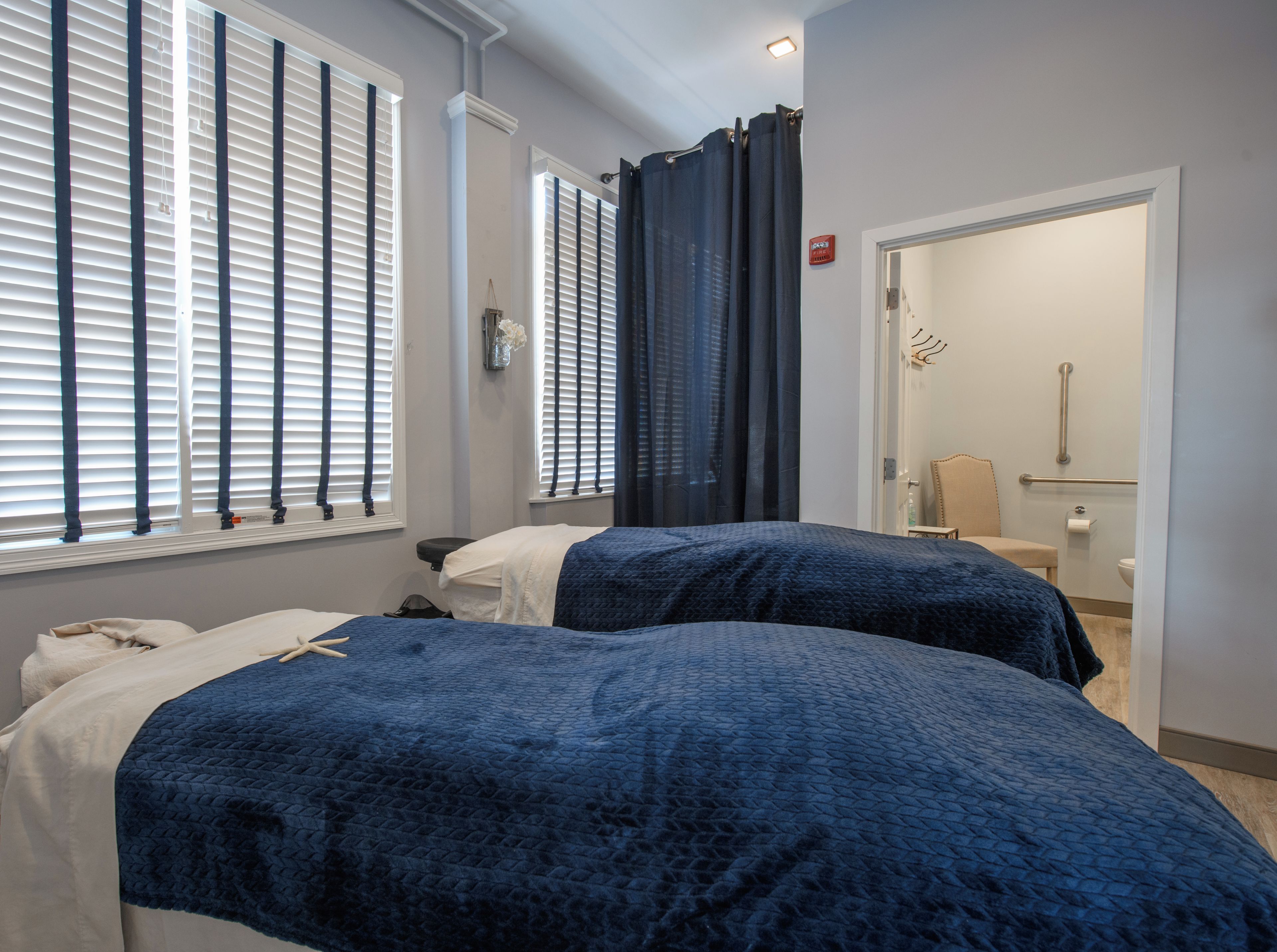 A hotel room with two beds, a bathroom with a toilet and handrail, and a window with blinds.