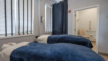 A hotel room with two beds, a bathroom with a toilet and handrail, and a window with blinds.
