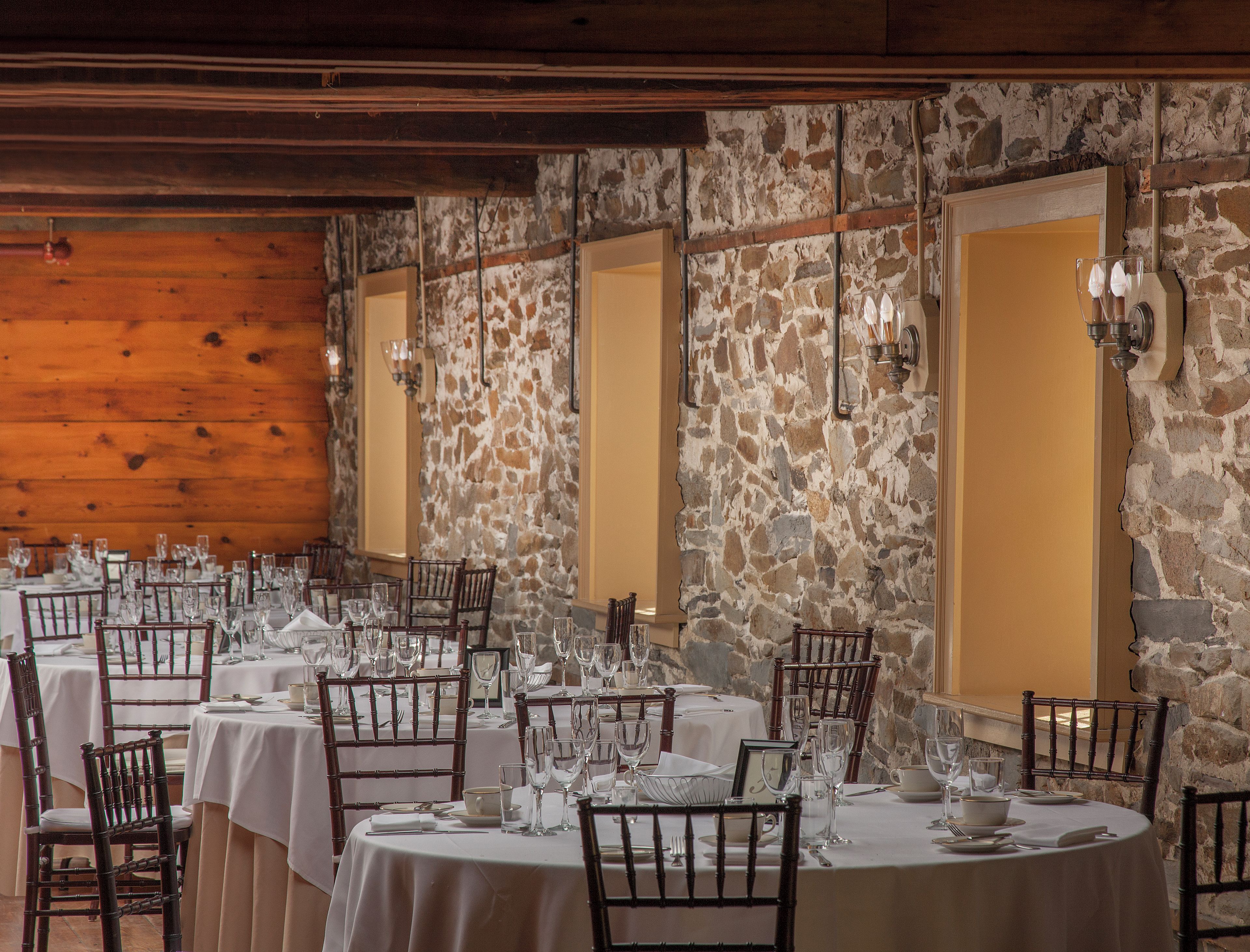 A dining area with round tables set for a meal, wooden chairs, and stone walls.