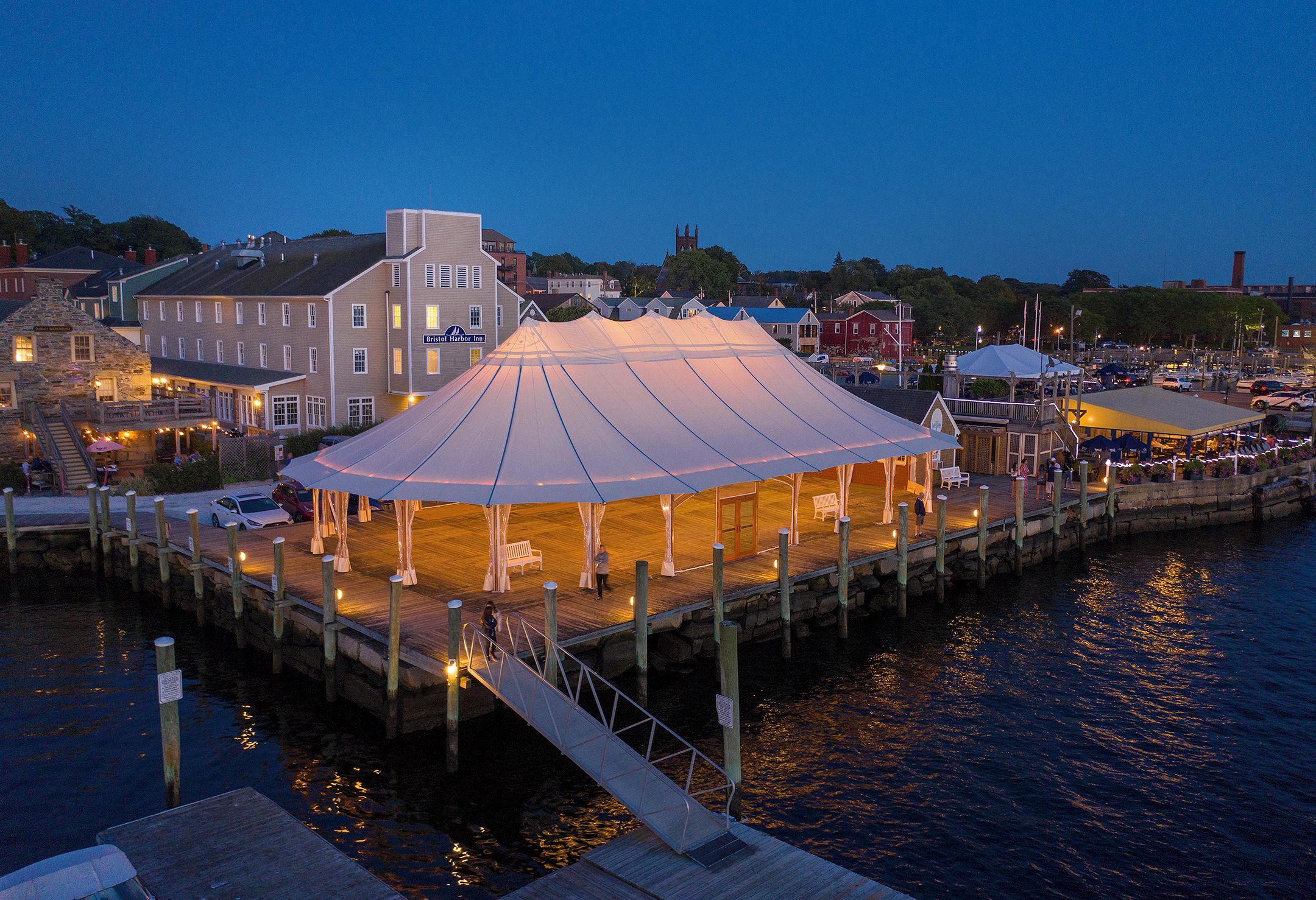 A waterfront pavilion with a covered seating area, a dock, and a view of the town and a church in the background.