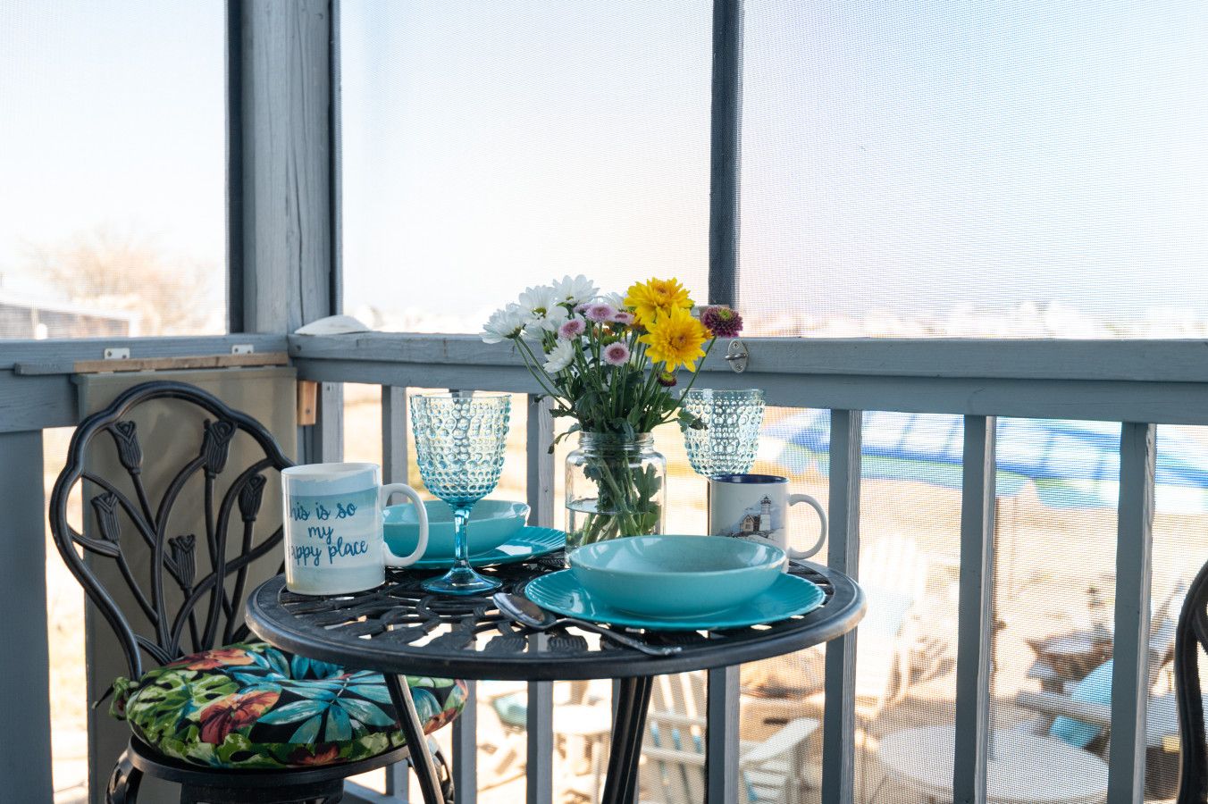 A balcony with a table set for two, featuring a vase with flowers, a cup with a message, and a decorative plate.