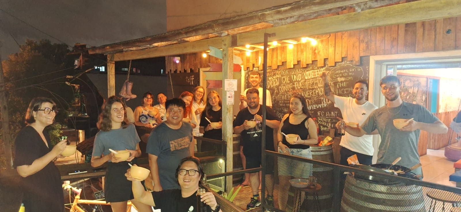 Un grupo de personas cenando en un puesto de comida al aire libre con una estructura de madera rústica y un menú en una pizarra.