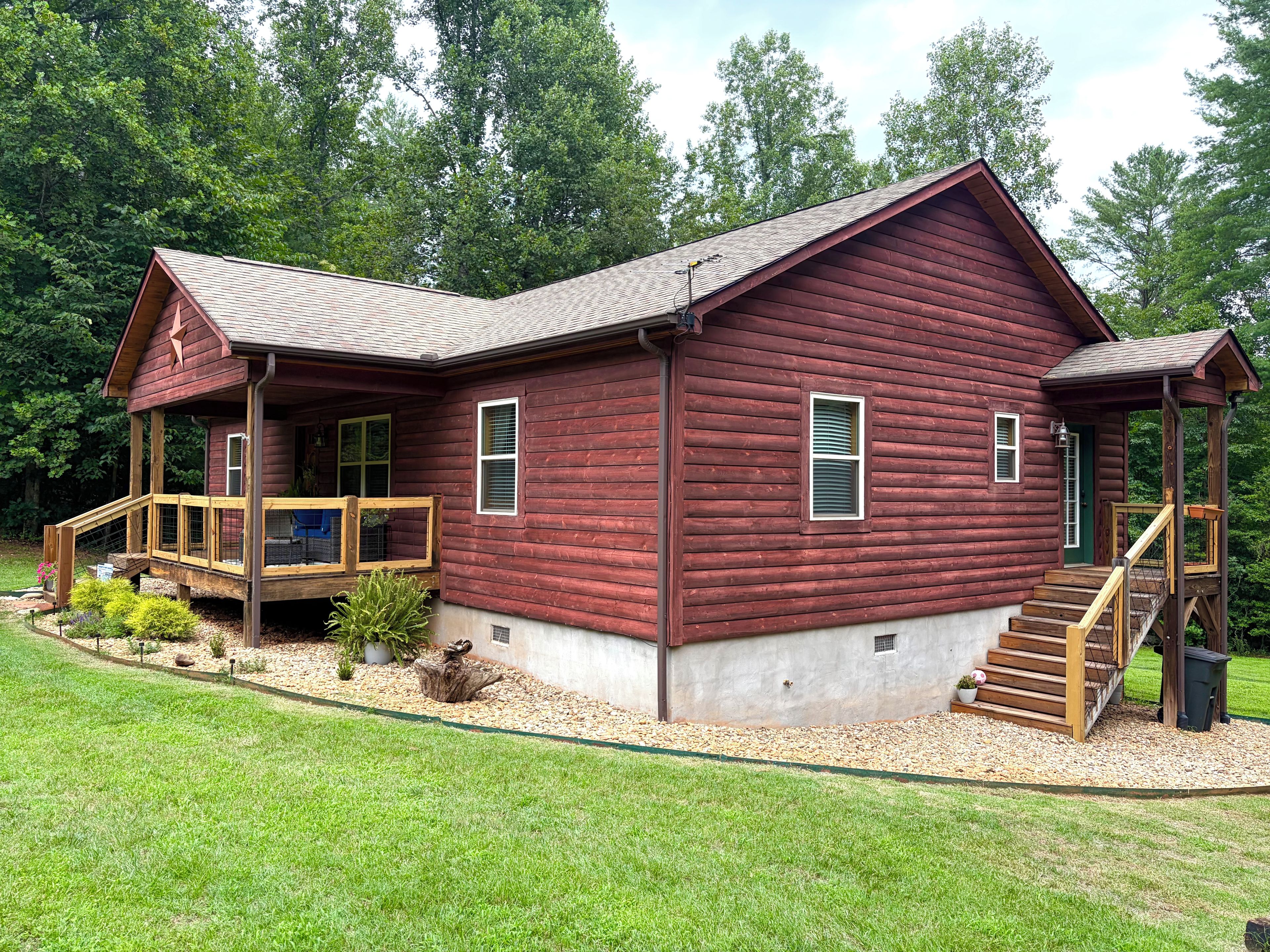 A two-story log cabin with a porch and a small deck, surrounded by a well-maintained lawn and trees.