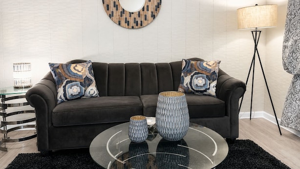 Ein modernes Wohnzimmer mit einem dunkelbraunen Sofa, gemusterten Zierkissen, einem runden Glastisch und einem wandmontierten Spiegel.