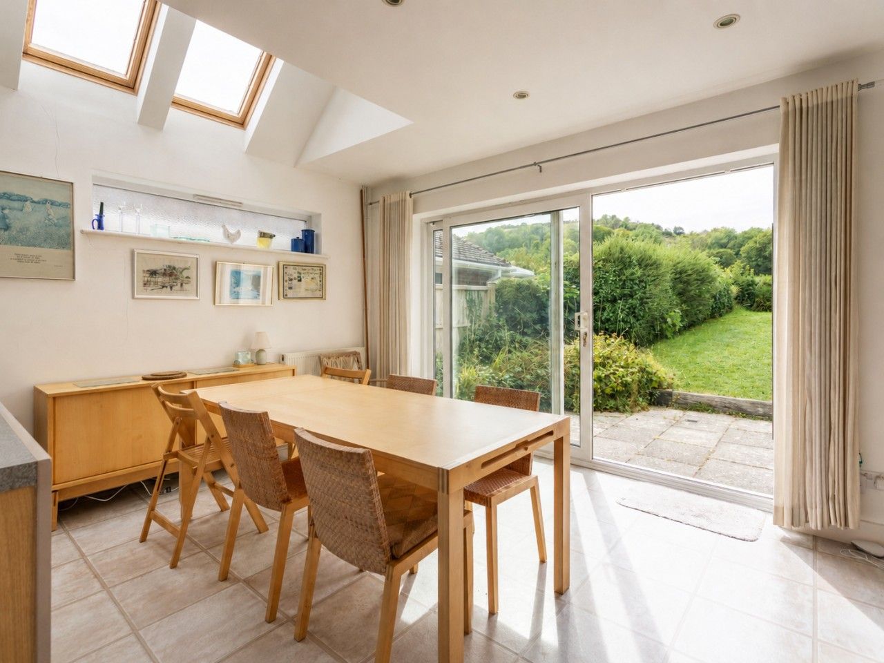 A dining area with a large table and chairs, a wooden cabinet, and a view of a garden through a sliding glass door.