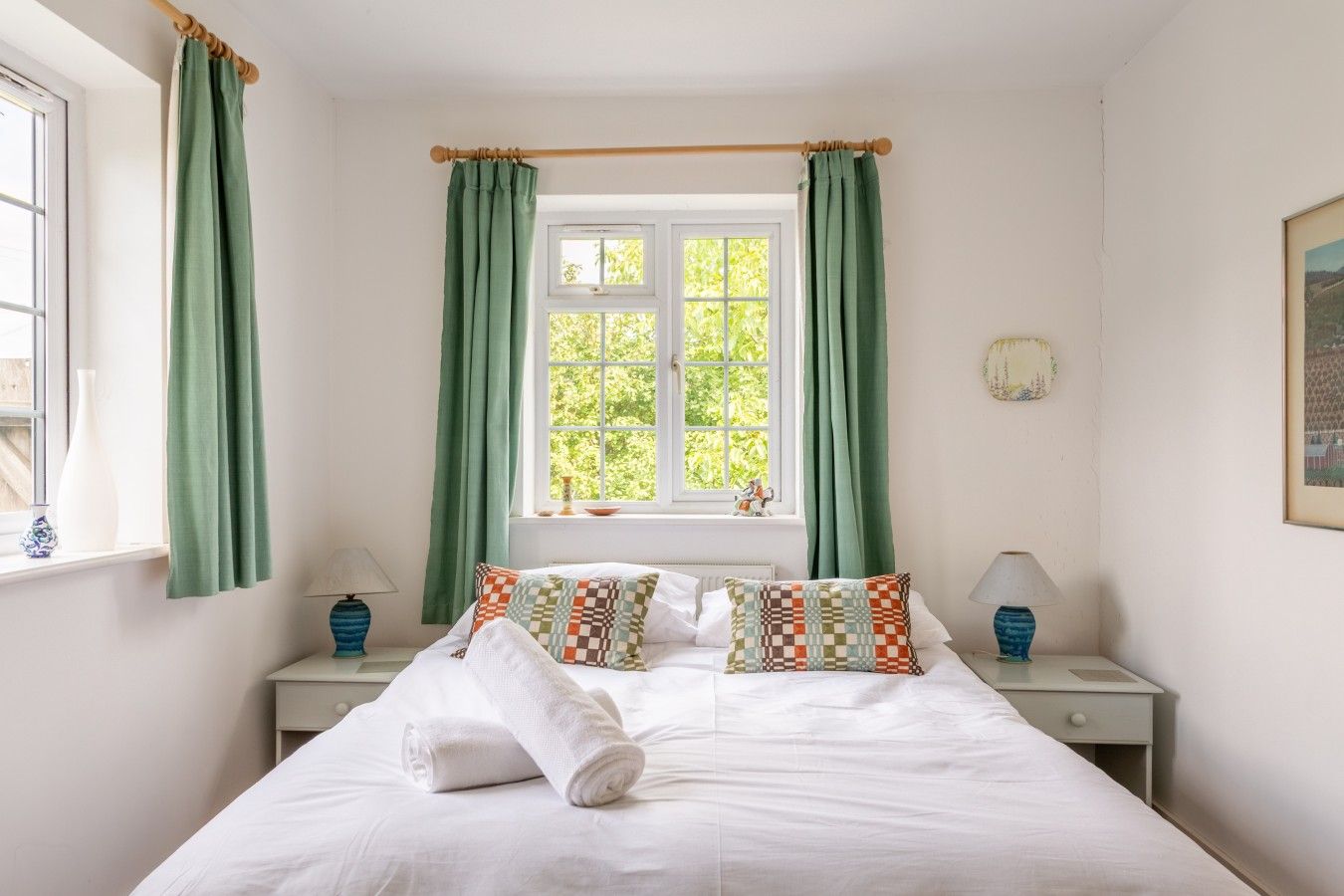 Une chambre à coucher avec un lit, deux tables de chevet, une fenêtre avec des rideaux et une vue sur la verdure à l’extérieur.