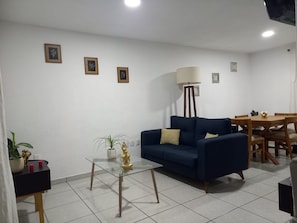 Ein Wohnzimmer mit einem blauen Sofa, einem Glastisch, einer Stehlampe und einem Esstisch mit Stühlen.