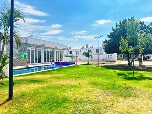Ein Poolbereich mit blauem Himmel, ein Gebäude mit großen Fenstern und ein von Bäumen gesäumter, eingezäunter Bereich.