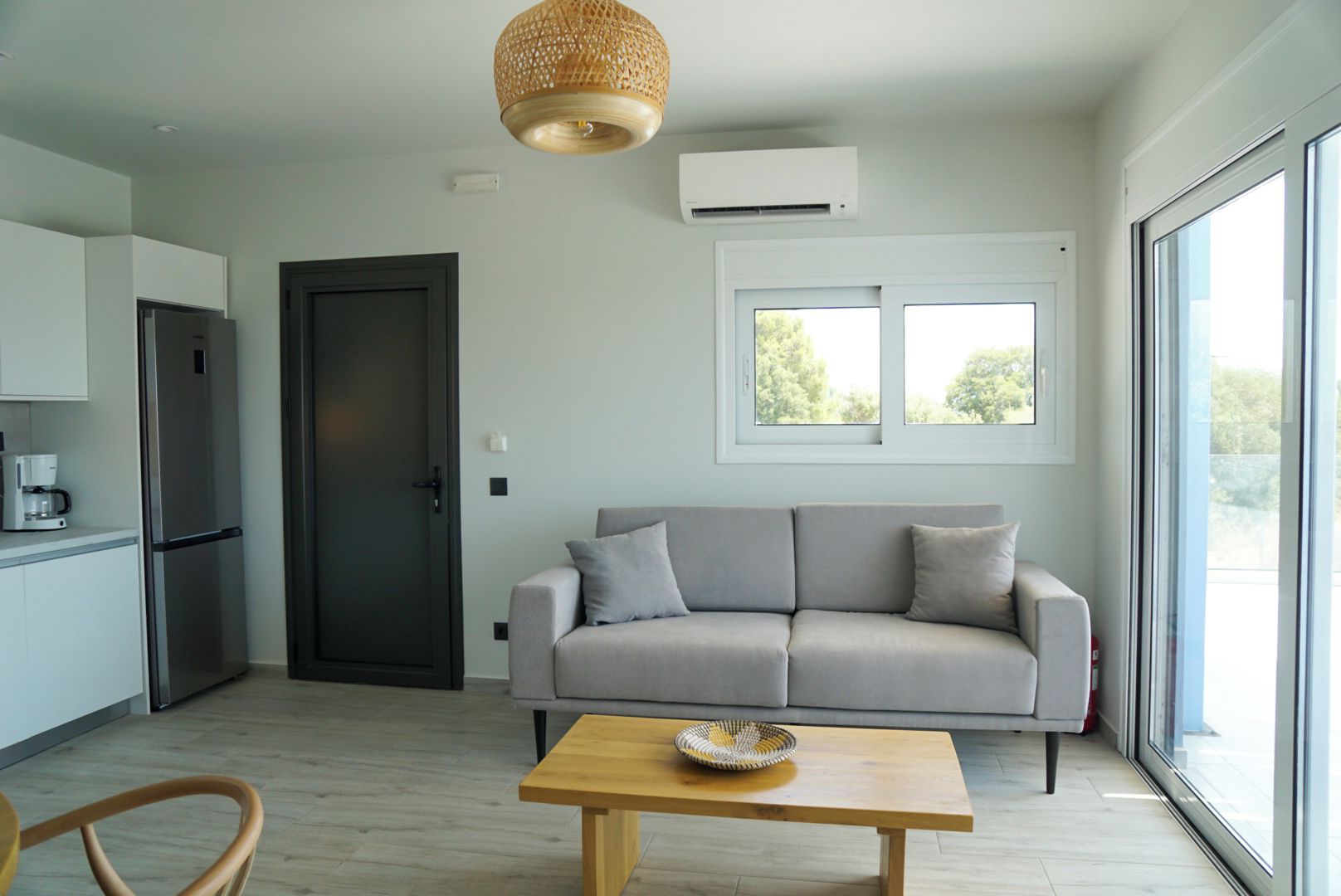 A modern living space with a sofa, a coffee table, a kitchen area with a refrigerator, and a large window providing natural light.