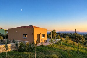 Une maison moderne avec un mur en pierre, une porte et des fenêtres, entourée de verdure et sous un ciel dégagé.