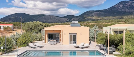 Una villa moderna con piscina, mobiliario de exterior y montañas al fondo.