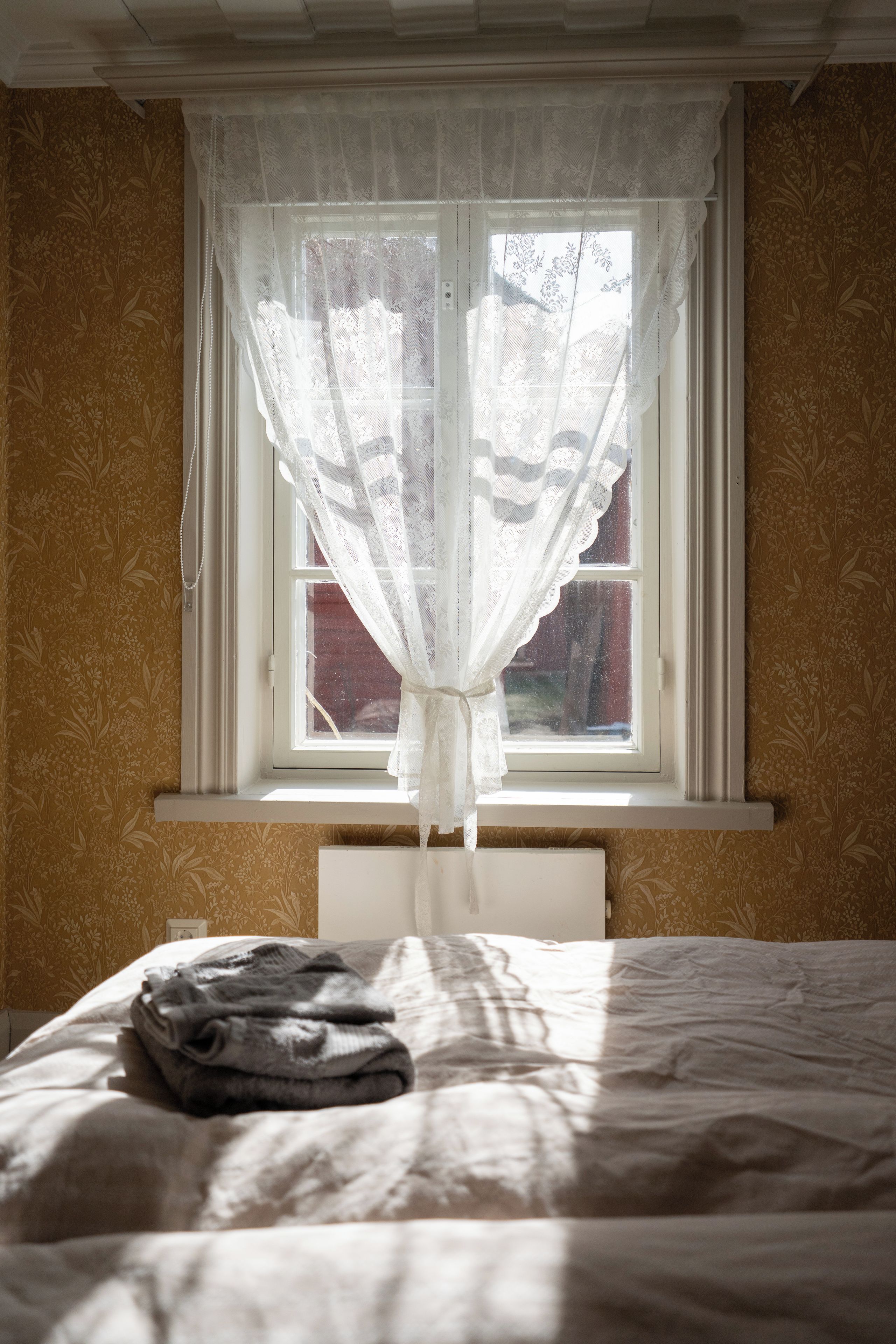 Un dormitorio con una cama, una ventana con cortina de encaje y una pared con estampado floral.