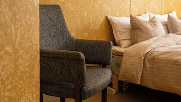 Un dormitorio con papel tapiz estampado, un sillón tapizado en gris, una cama con ropa de cama blanca y una mesita de noche.