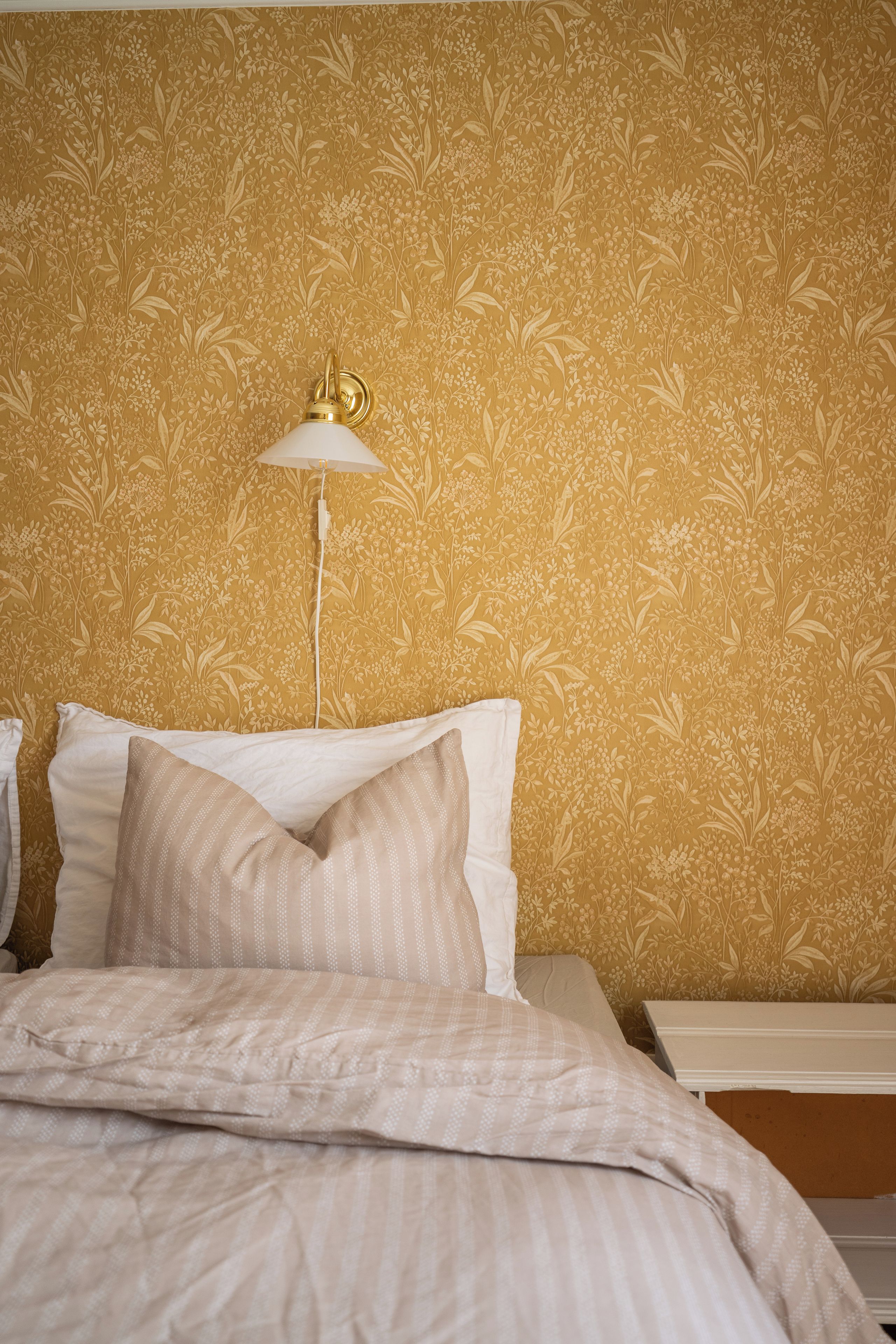 A bedroom with a floral wallpaper pattern, a white bed with multiple pillows, and a bedside lamp.