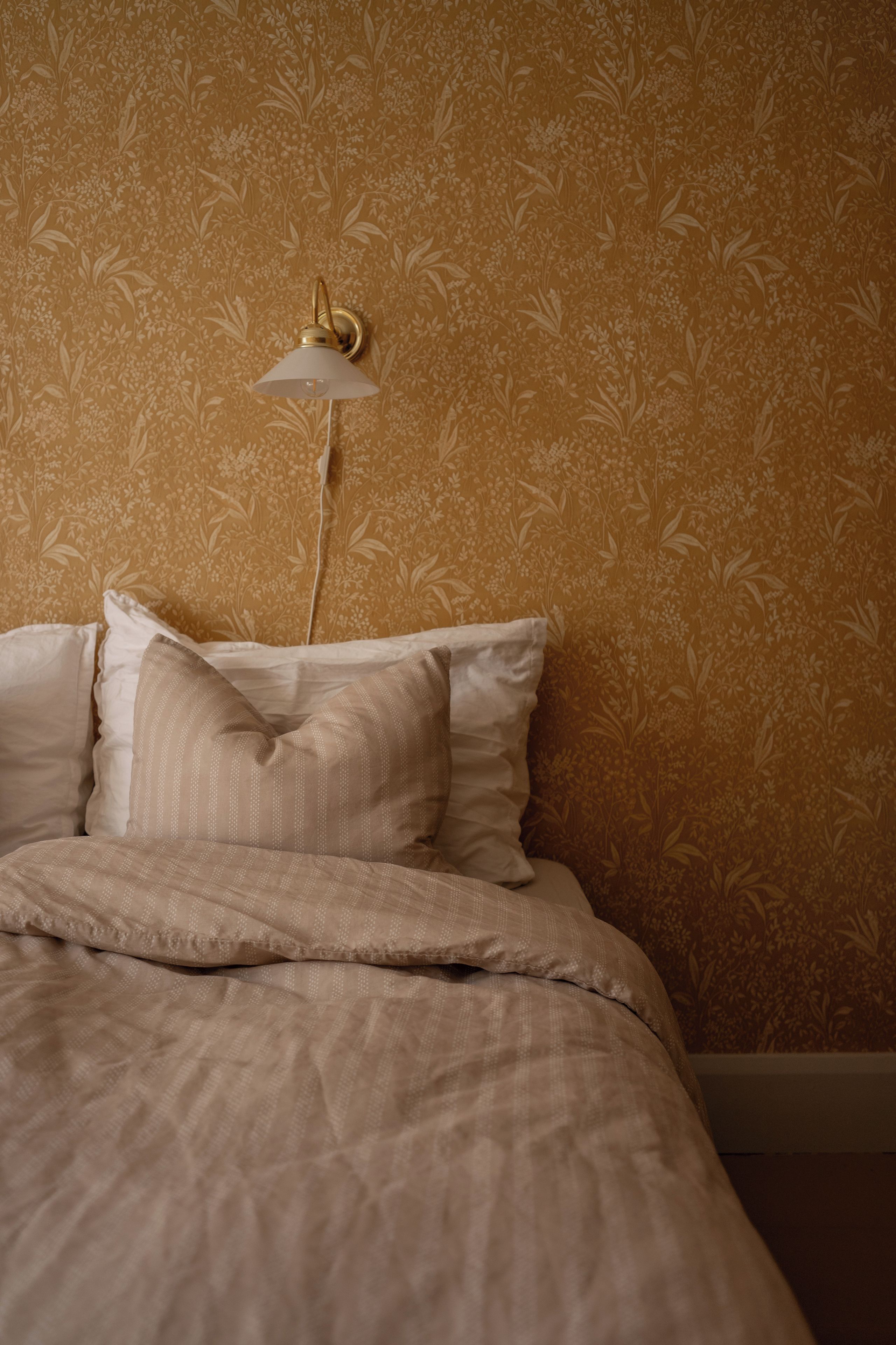 Una cama con un papel tapiz estampado y una lámpara fijada a la pared.