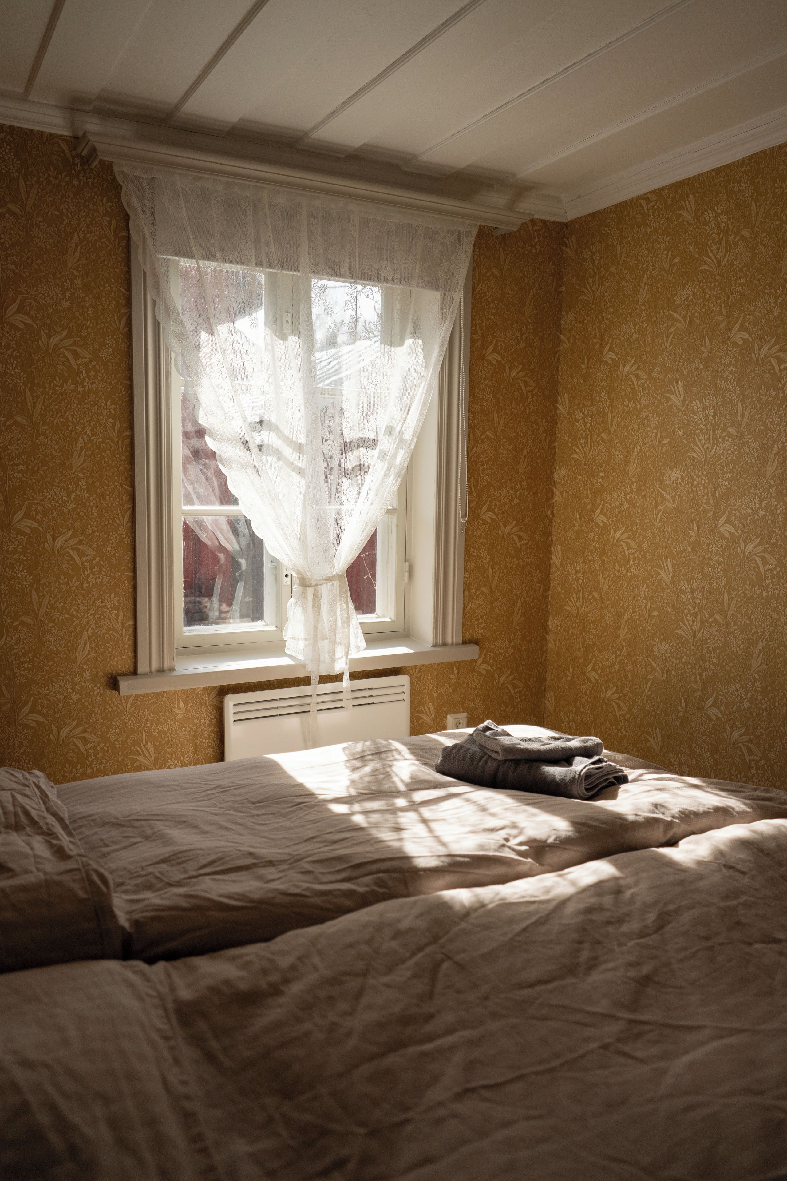 Quarto com cama, janela com cortina leve e radiador.