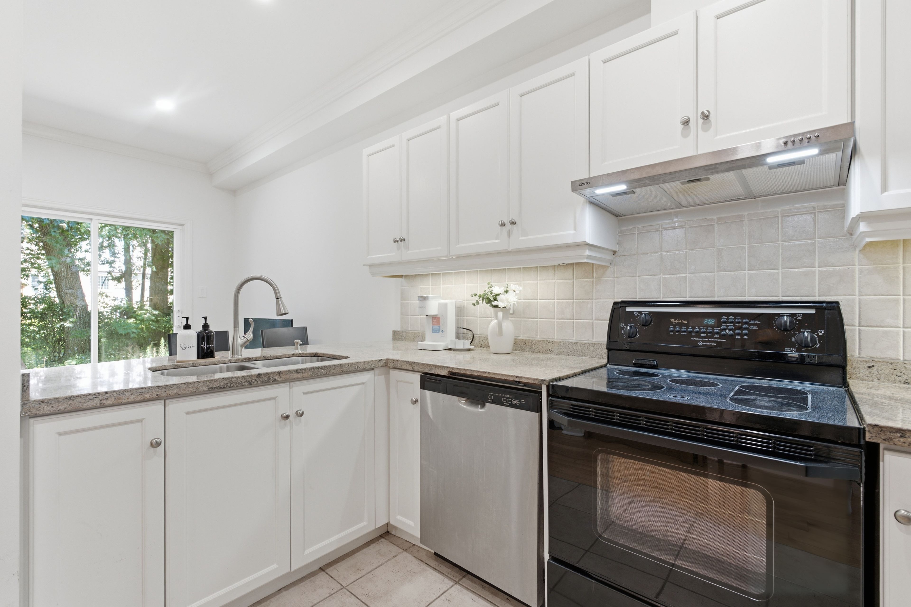 A modern kitchen with white cabinetry, a stainless steel dishwasher, a black stove, and a window with a view of trees.