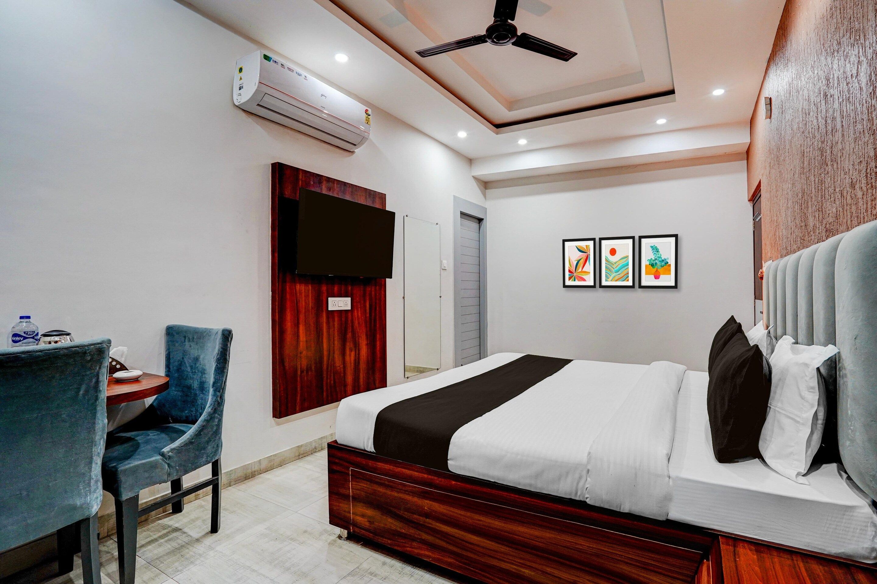 Een hotelkamer met een bed, een bureau, een stoel, een airconditioner, een plafondventilator en kunst aan de muur.