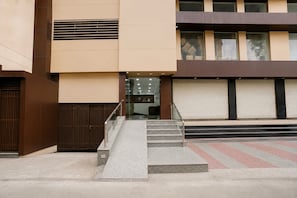 A building entrance with a set of stairs leading to a glass door, a metal handrail, and a closed shuttered storefront.