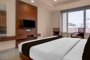 A hotel room with a bed, a television mounted on a wooden panel, a window with a view of buildings, and two chairs.