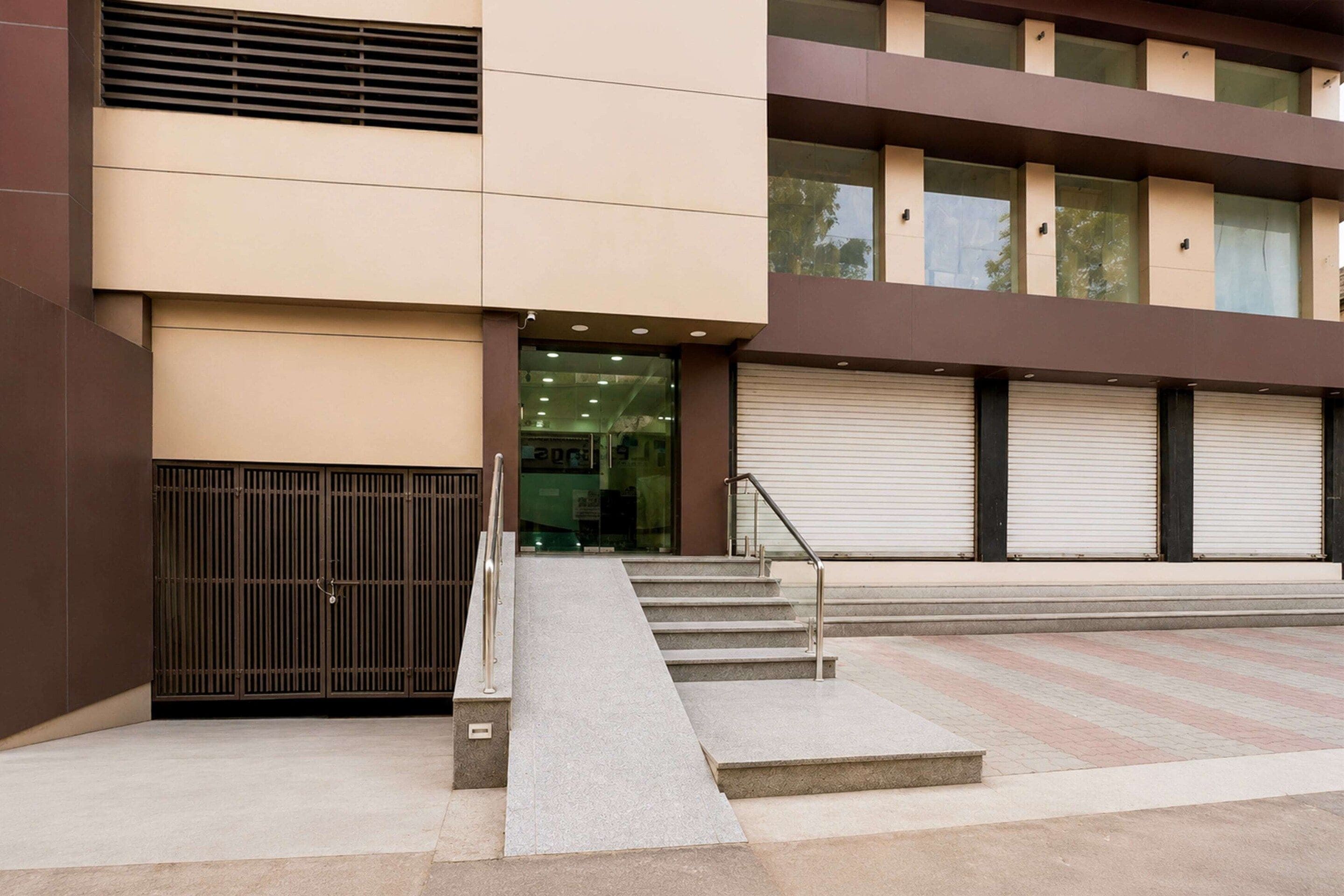 A building with a modern design, featuring a set of stairs leading to a glass entrance, a metal gate, and a series of closed roller shutters.