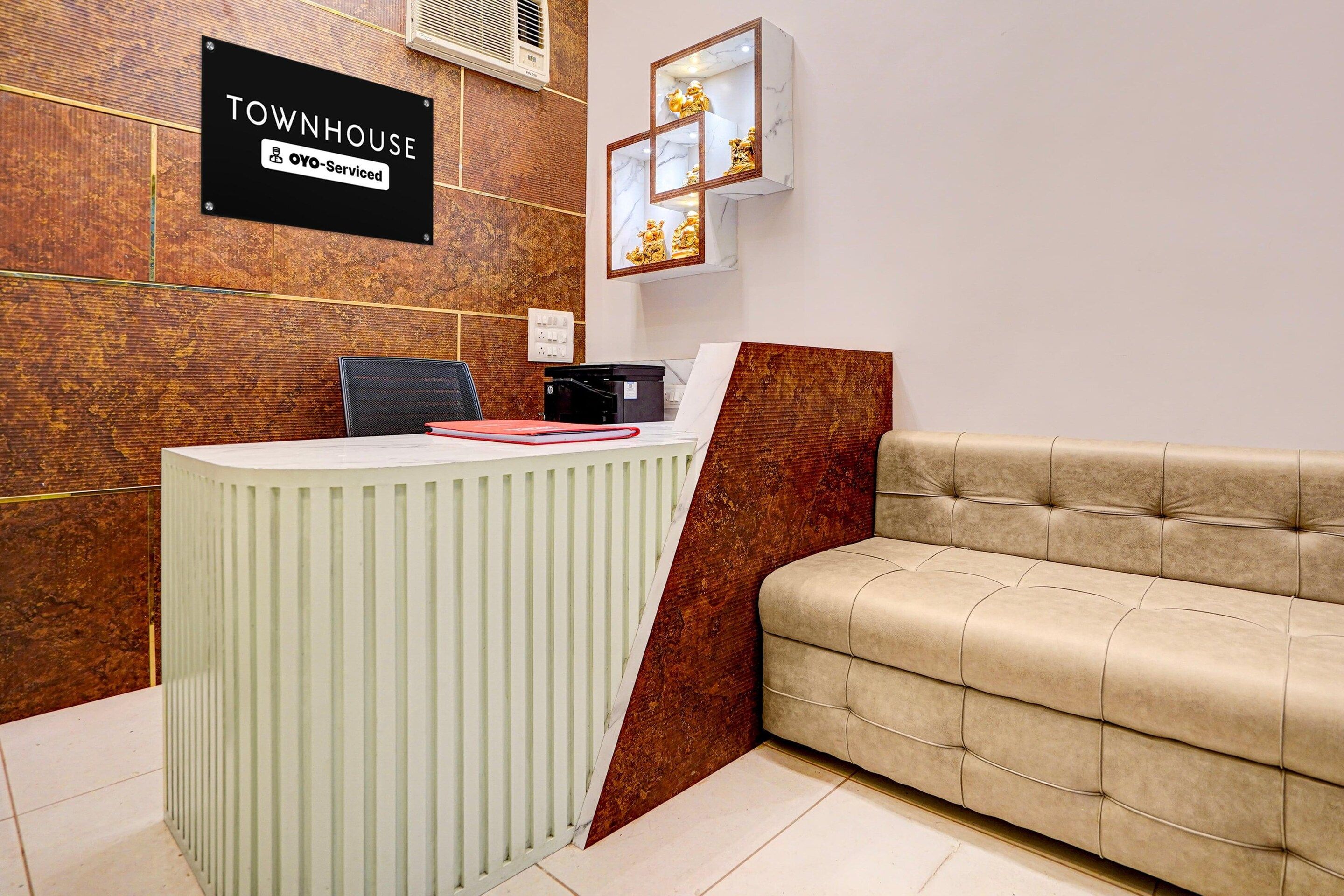 A reception desk with a sign indicating it is serviced by OYO, a reception area with a brown textured wall, a white cushioned sofa, and a marble-framed mirror.