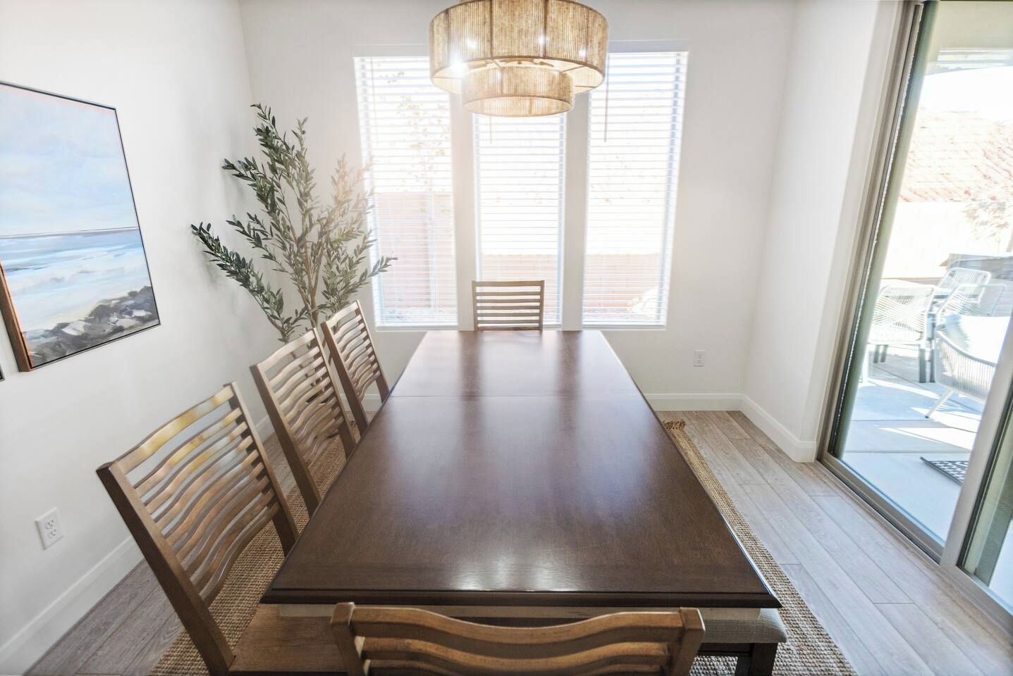 Een eetkamer met een lange tafel, houten stoelen, een hanglamp en een groot raam met jaloezieën.