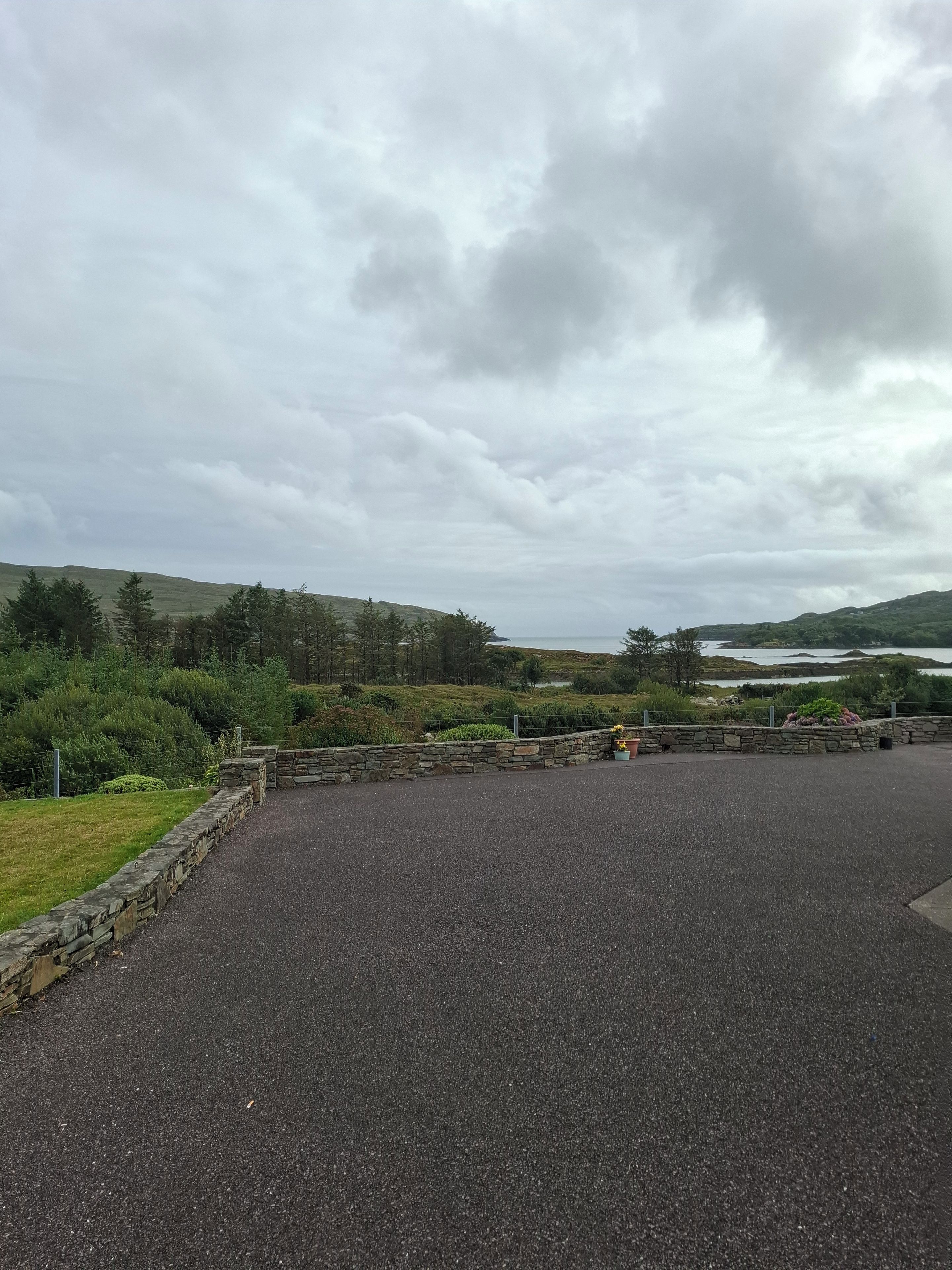 A paved area with stone walls and greenery, leading to a body of water and hills in the distance.