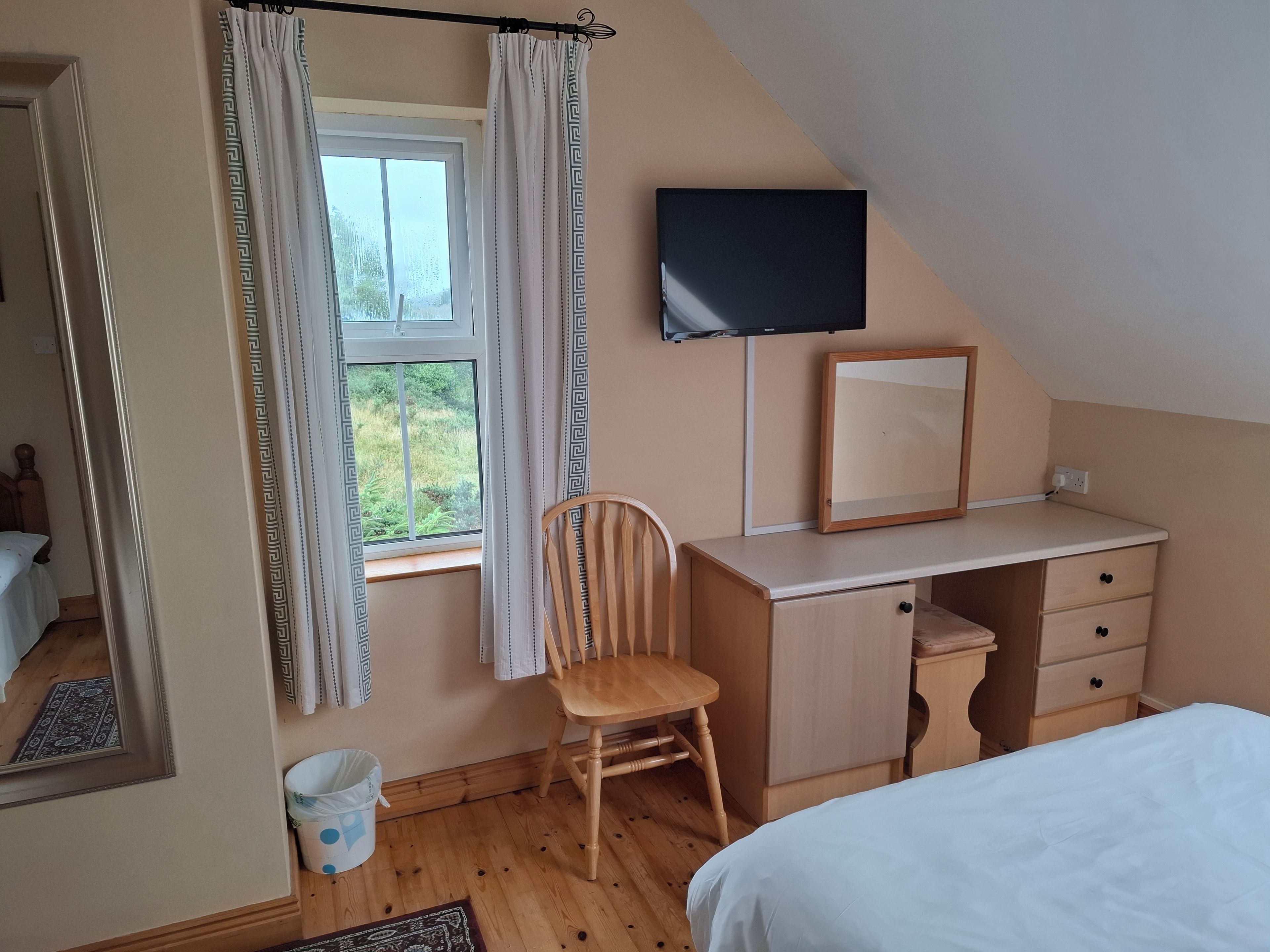 Un dormitorio con cama, escritorio, silla, televisor y ventana con cortinas.