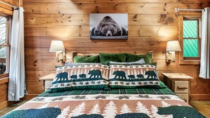 Une chambre à coucher dans un chalet en bois, avec un lit, deux lampes de chevet, un oreiller vert et une couverture à motifs d’ours.