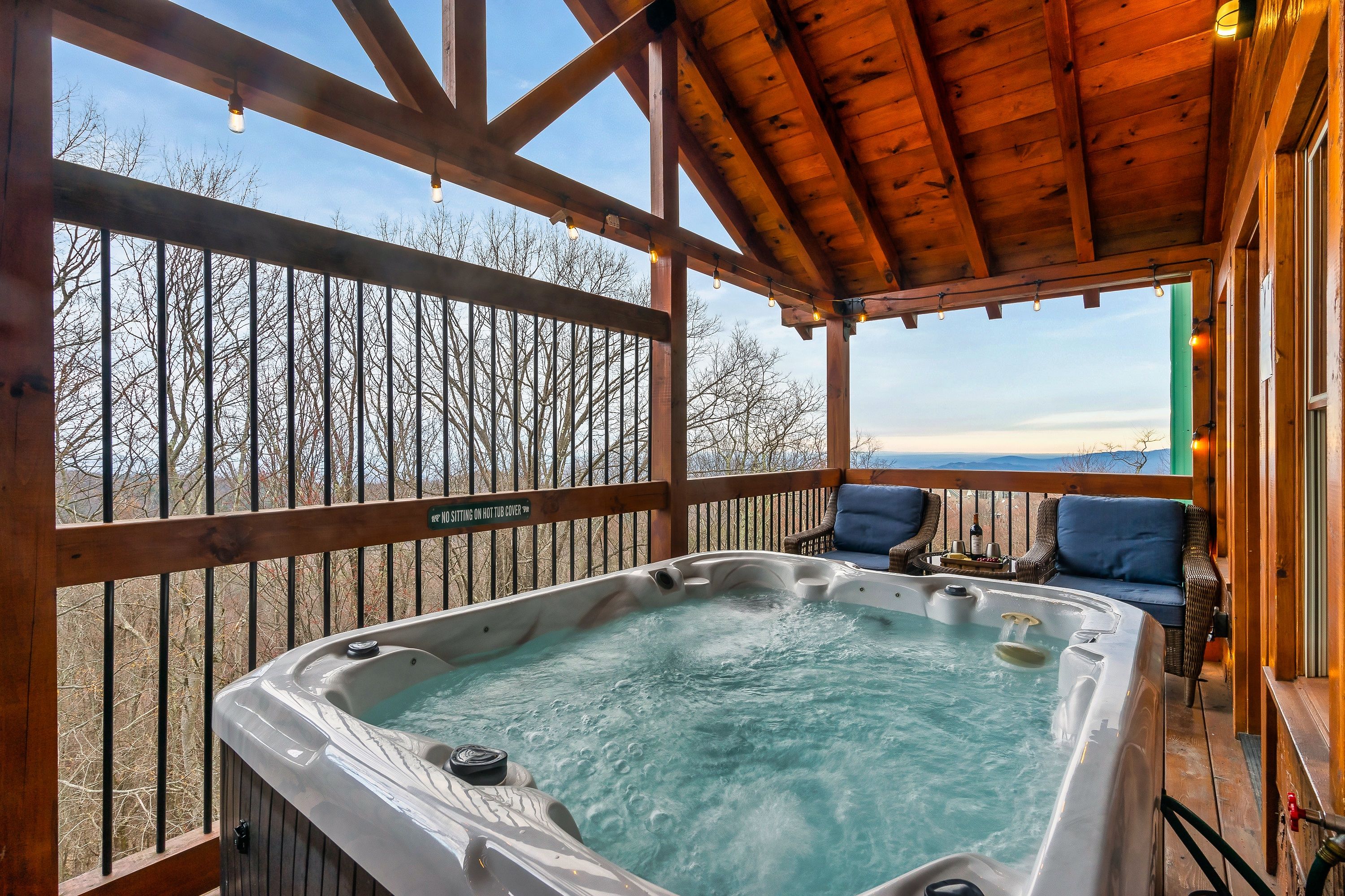 A hot tub on a wooden deck with chairs, a view of trees, and a sign that says "STIRRING ON HOT TUBS."