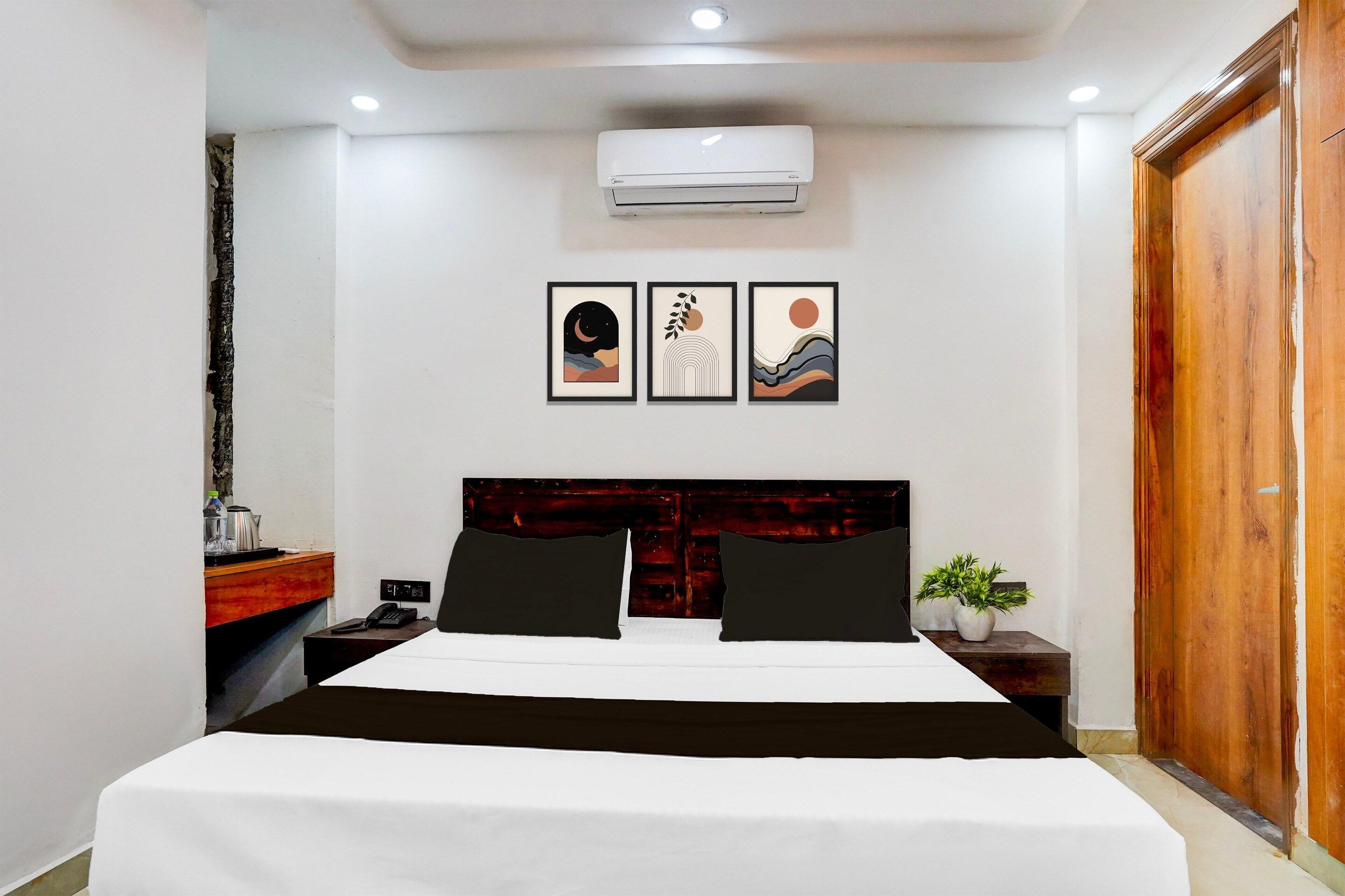 Habitación de hotel con cama, mesita de noche, teléfono, planta y cuadros en la pared.