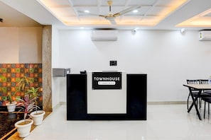 A reception desk with a sign indicating it is serviced by a company. There are potted plants, a ceiling fan, and a table with chairs to the right. - Premium Townhouse Sector 143b (Noida)